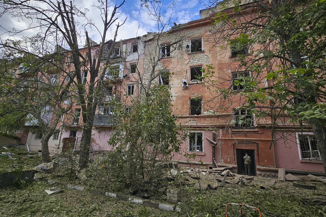 A destroyed apartment building in Kherson, Ukraine, is visible through a line of trees. The walls of the building are rose pink and terracotta, the windows have been blown out and debris is littered around the building.