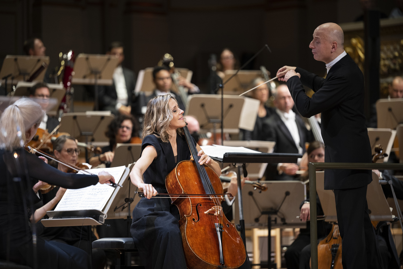 Sol Gabetta plays the cello at the forefront of the image, with an orchestra and music stands around and behind her and a male conductor, wearing a black suit and white shirt, to her right. She wears a navy dress, has long blonde curly hair and is smiling with her eyes closed while playing.