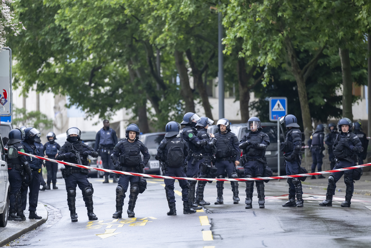 a large number of police in riot gear stand across a wet street with "do not cross" tape in front of them and green trees behind