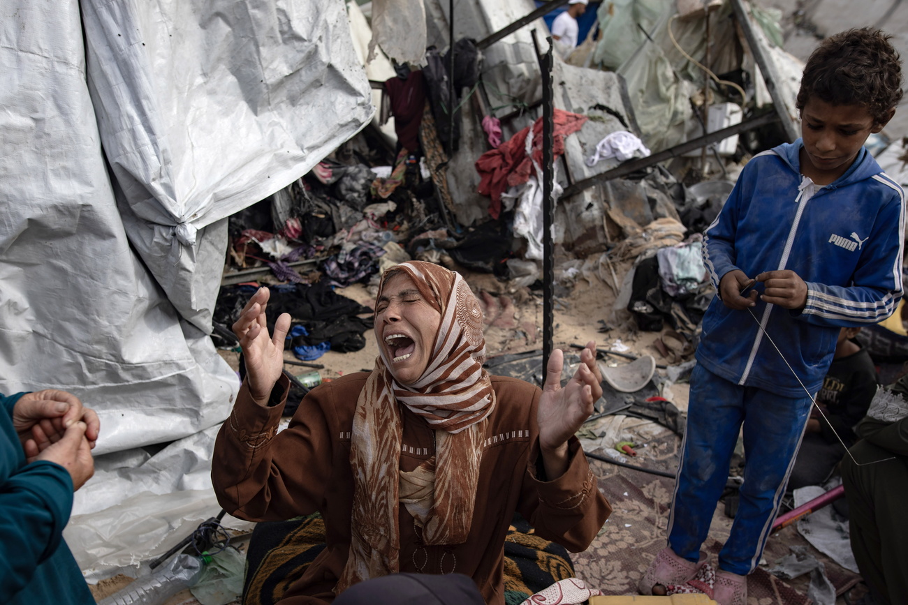 A woman in a brown top and brown and white headscarf is on her knees, screaming, amidst the wreckage of her tent. A child in a white striped blue Puma tracksuit is standing beside her.