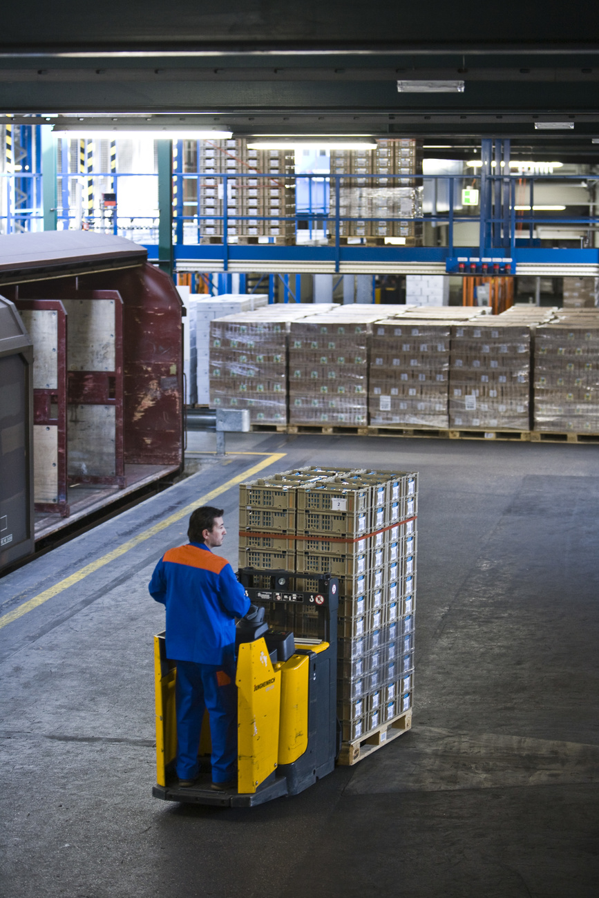 A man in a warehouse rides on an electric forklift with a number of boxes stacked on it.
