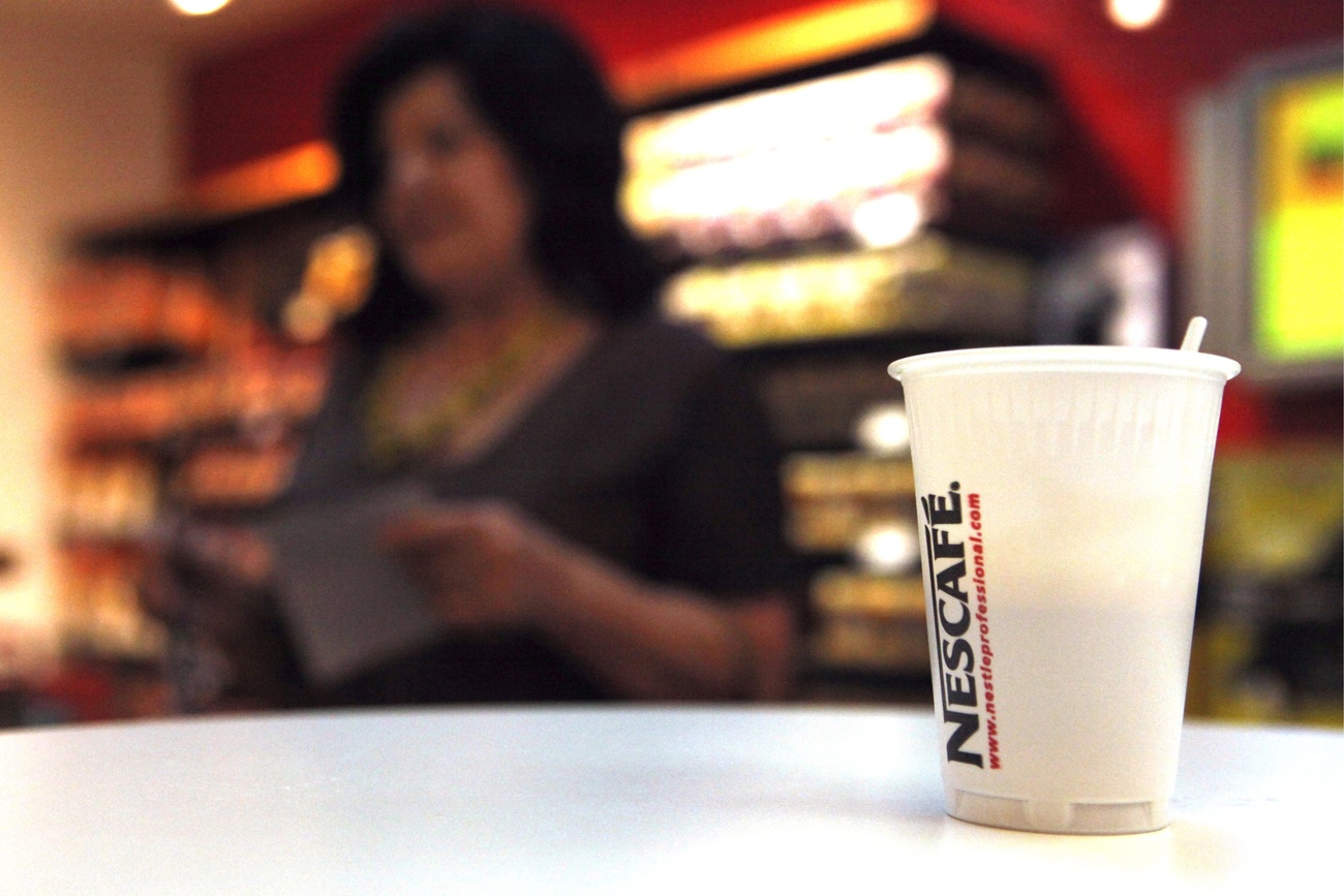 A plastic cup with the Nescafe logo sits on a countertop. Behind it, out of focus, is a shop employee.