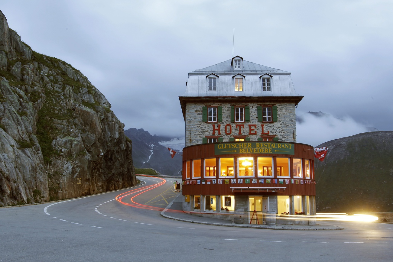A hotel with illuminated windows is shown on a mountain pass, with vehicle lights blurring by.