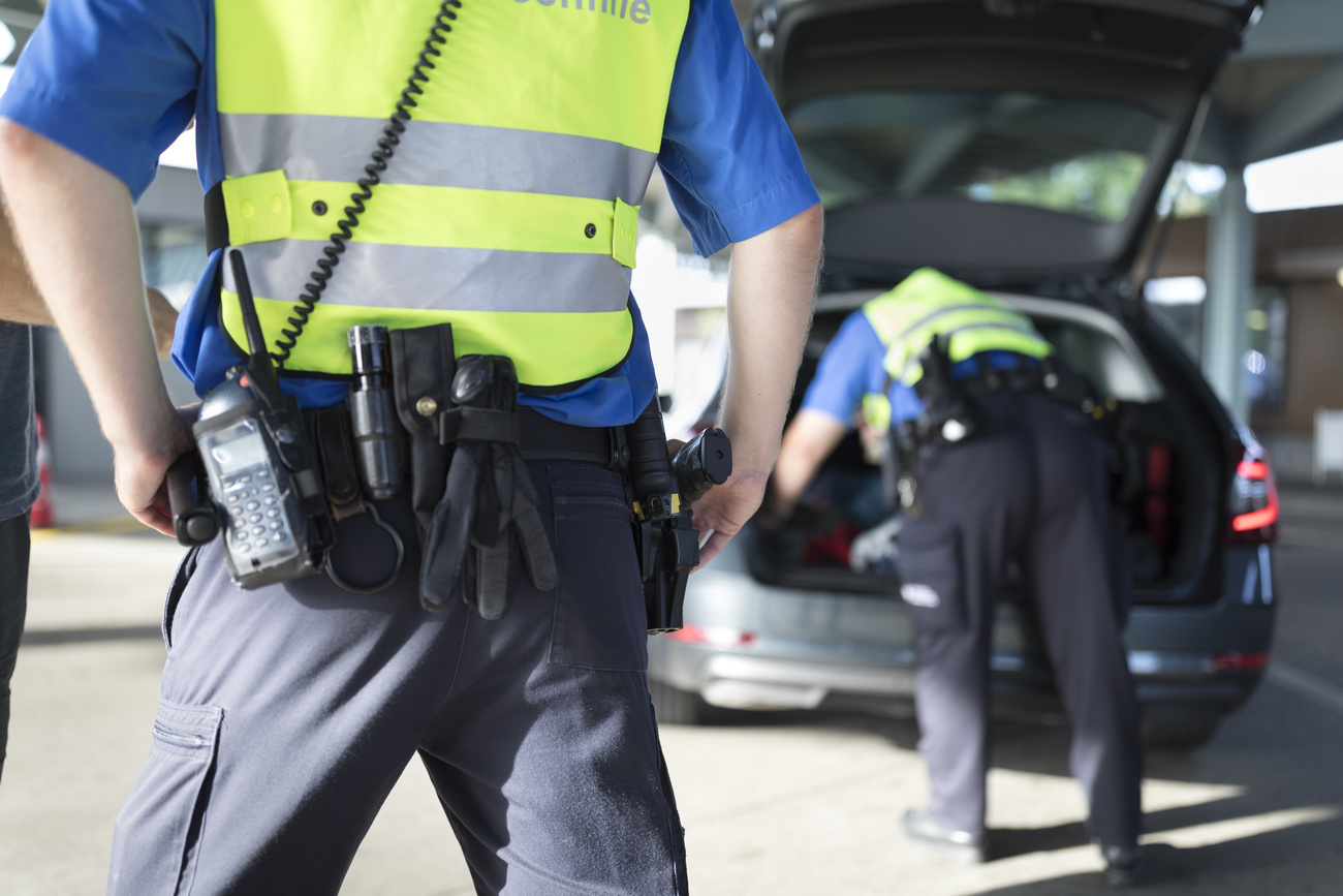 Two border guards are photographed from behind. They are wearing black trousers, blue shirts and high visibility vests, with black utility belts holding flashlights, gloves and walkie talkies. One stands with his hands on his hips in the foreground of the image, the other is leaning into the boot of a car.