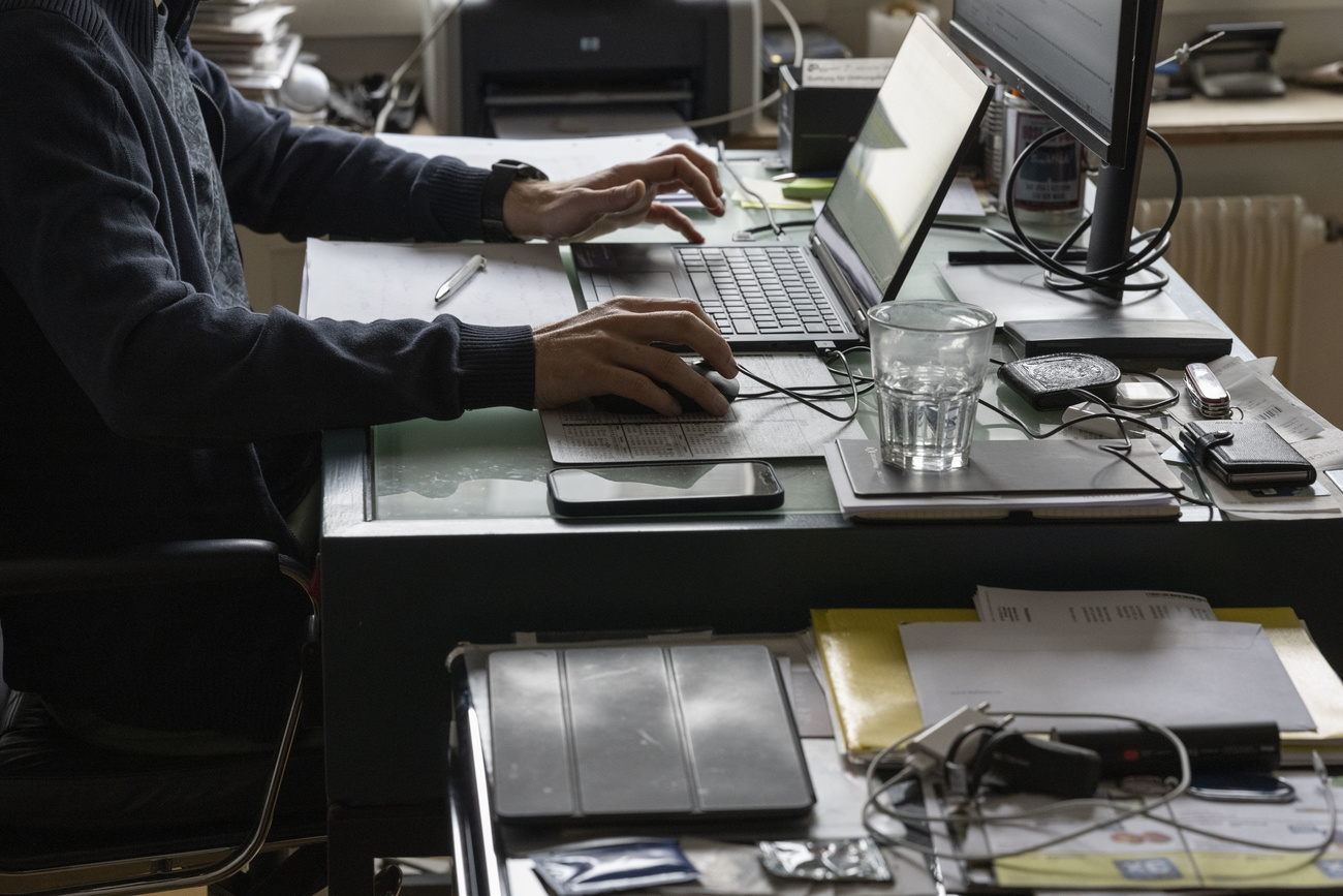 A man sitting at a desk wotking on his laptop. he is wearing a blue suit. beside him are various papers, stacked, cables and a glass of water.