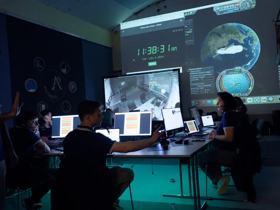 giovani seduti a delle scrivanie con dei monitori in quello che ricorda un centro di controllo