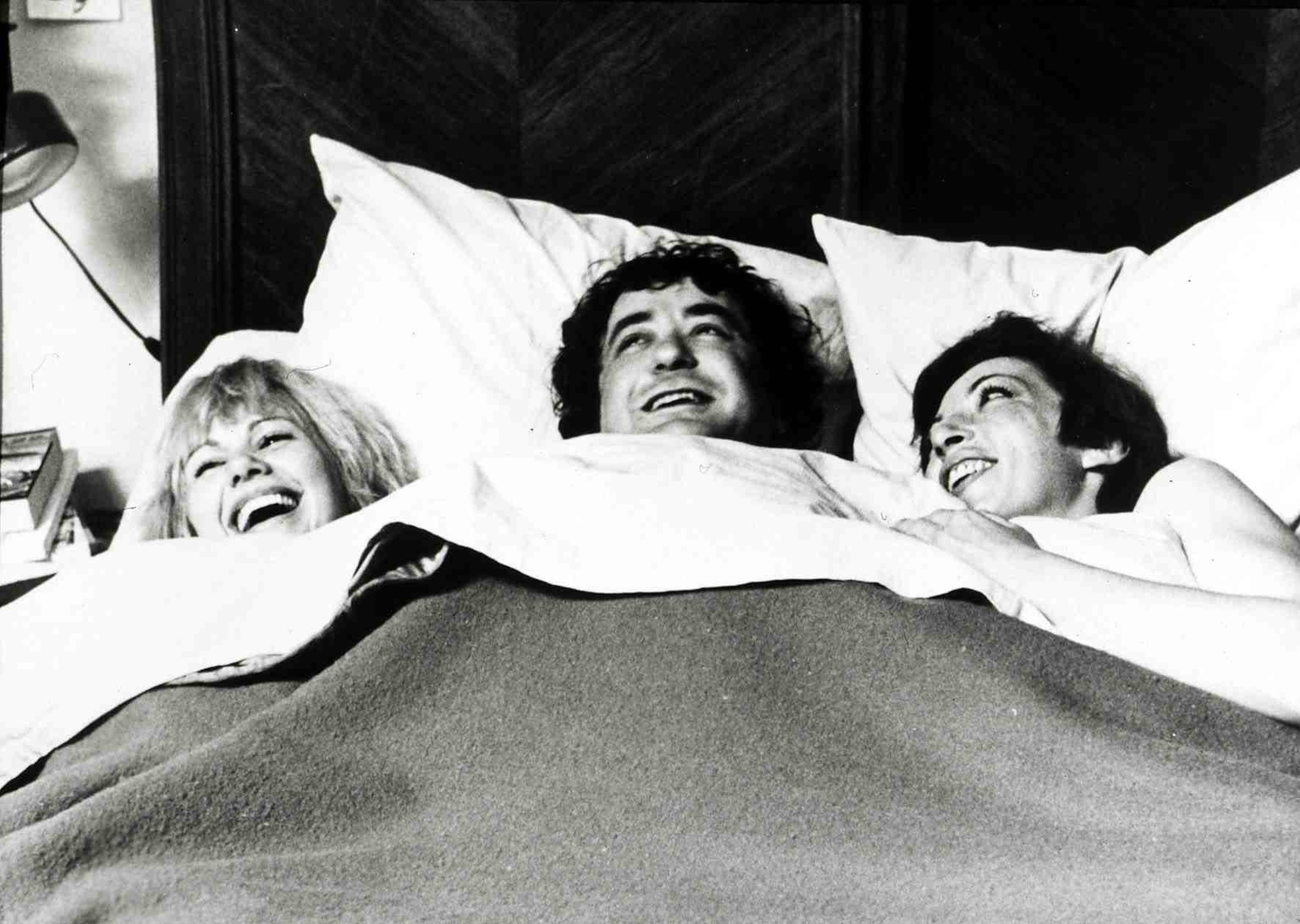 Ein Mann und zwei Frauen unter einer Bettdecke, alle lachen.