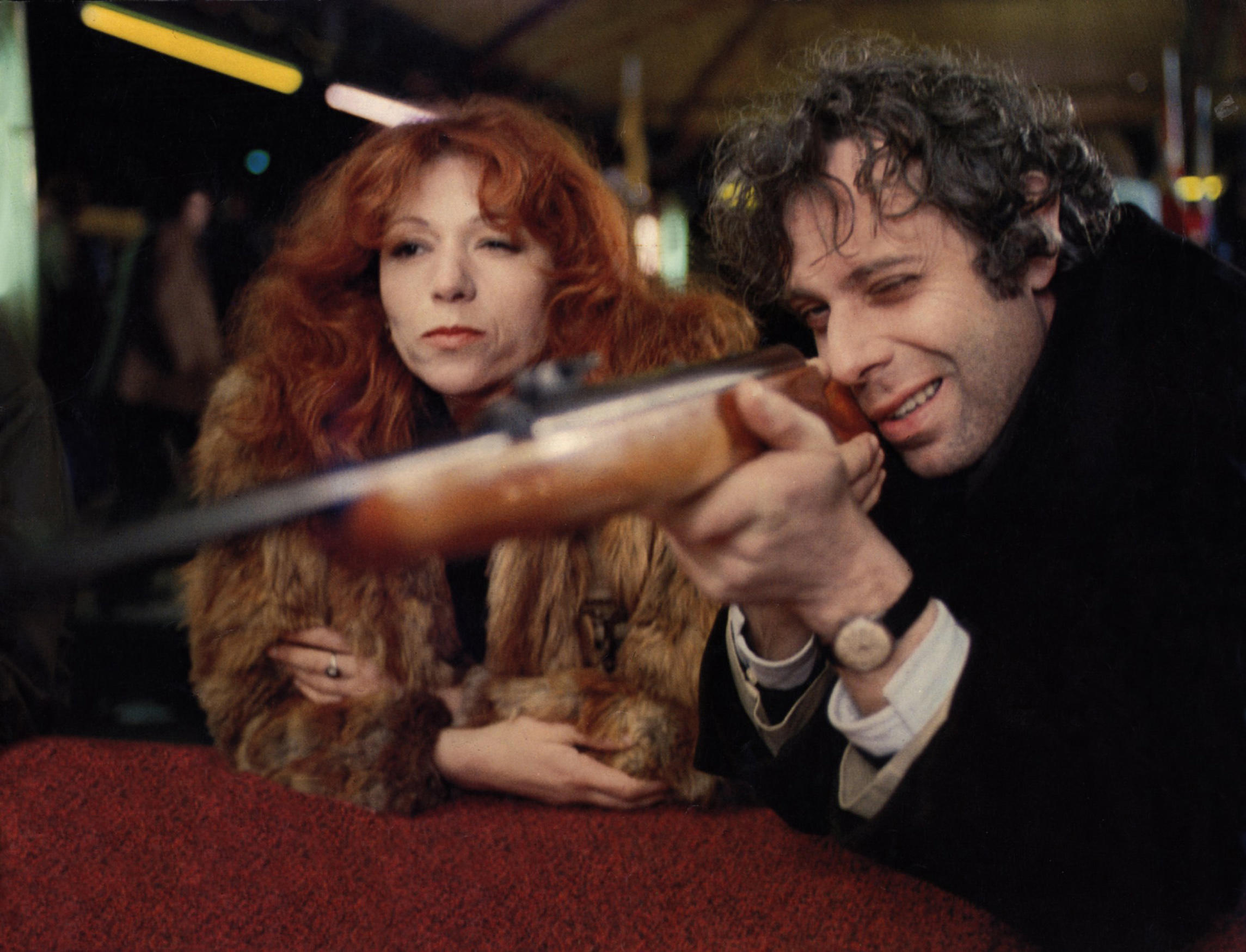 Ein Mann mit einem Gewehr an einer Schiessbude, daneben eine Frau im Pelzmantel.