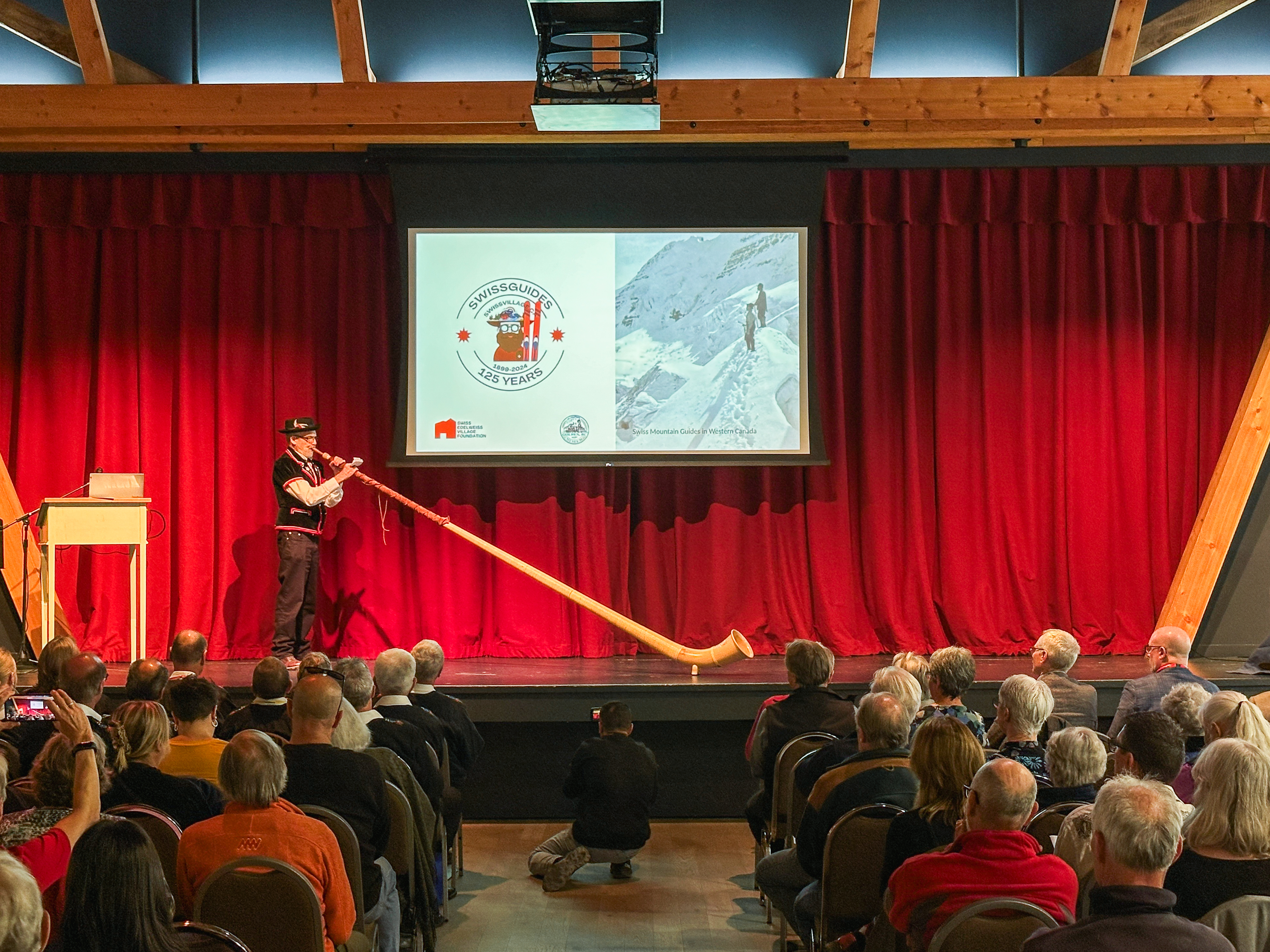 Homme jouant du cor des Alpes sur une scène devant un public assis.