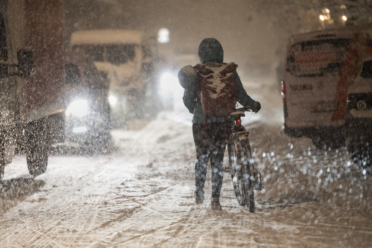 2024年11月21日星期四突降大雪,在瑞士伯尔尼的洛林区,一名骑行者推着自行车从被困在大雪中的汽车旁经过。