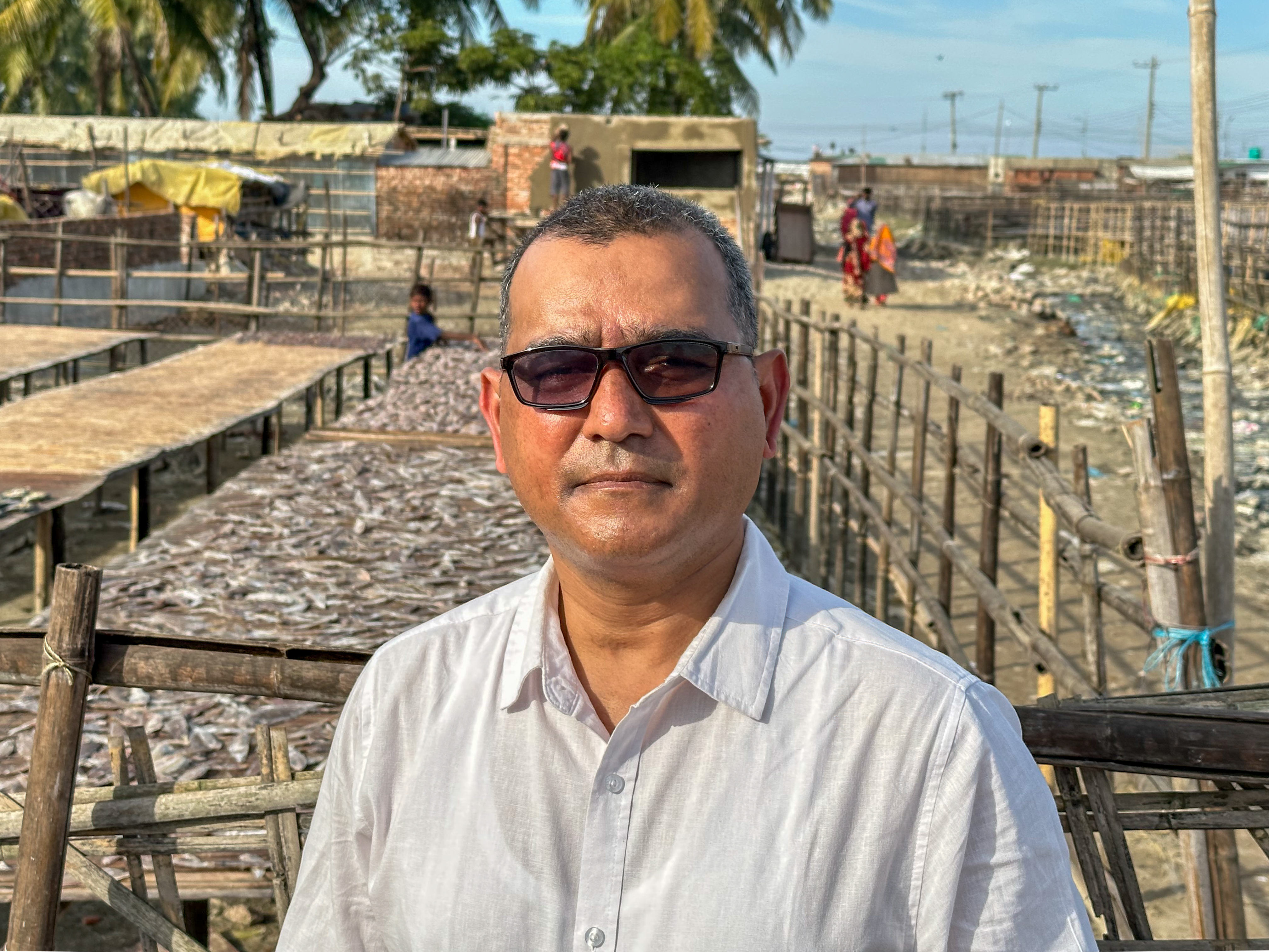 Person mit Sonnenbrille in weissem Hemd vor getrocknetem Fisch auf Gestellen.