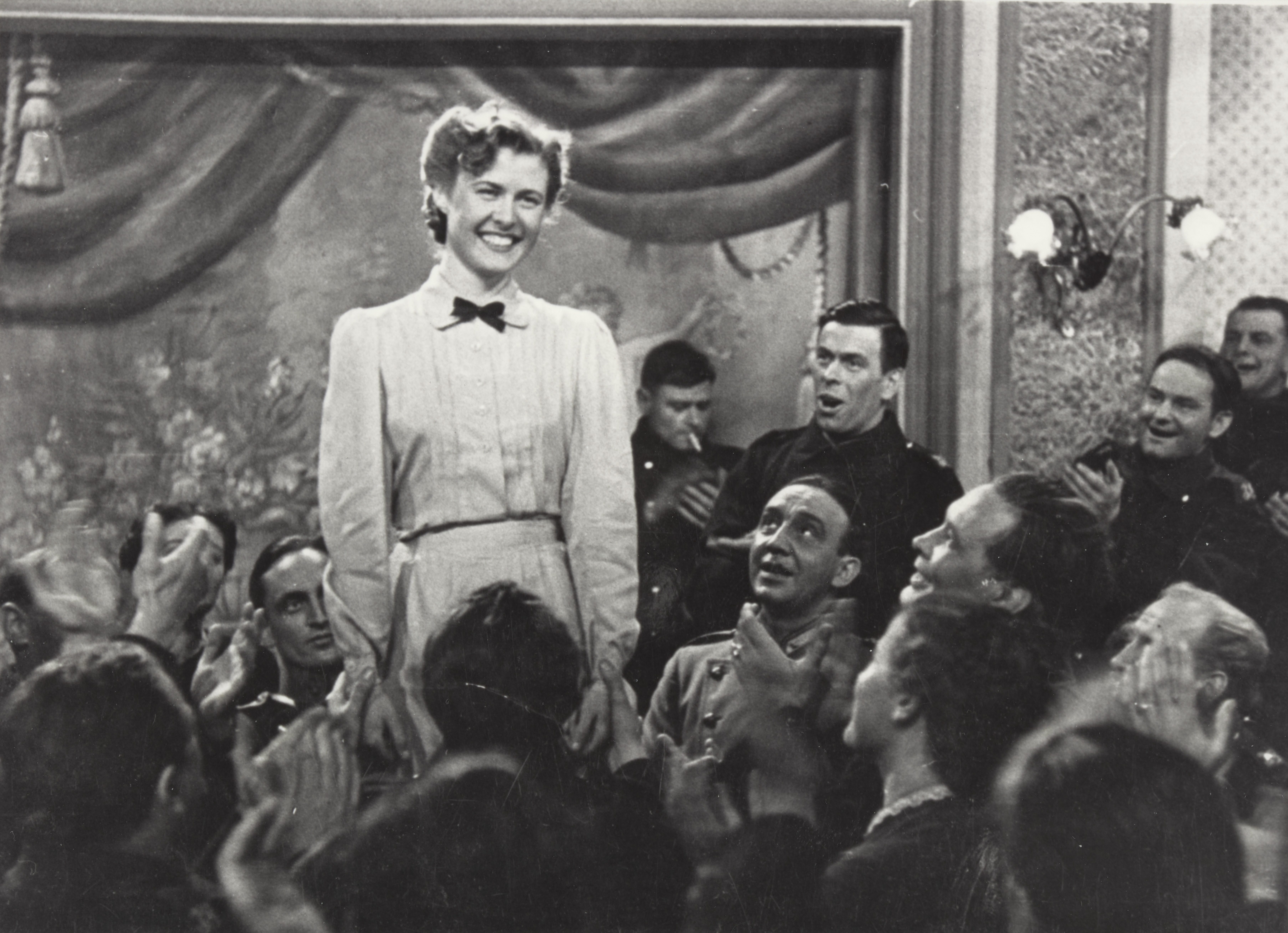 Black and white scene. A woman in a white dress stands in the centre of the stage, surrounded by an applauding crowd.