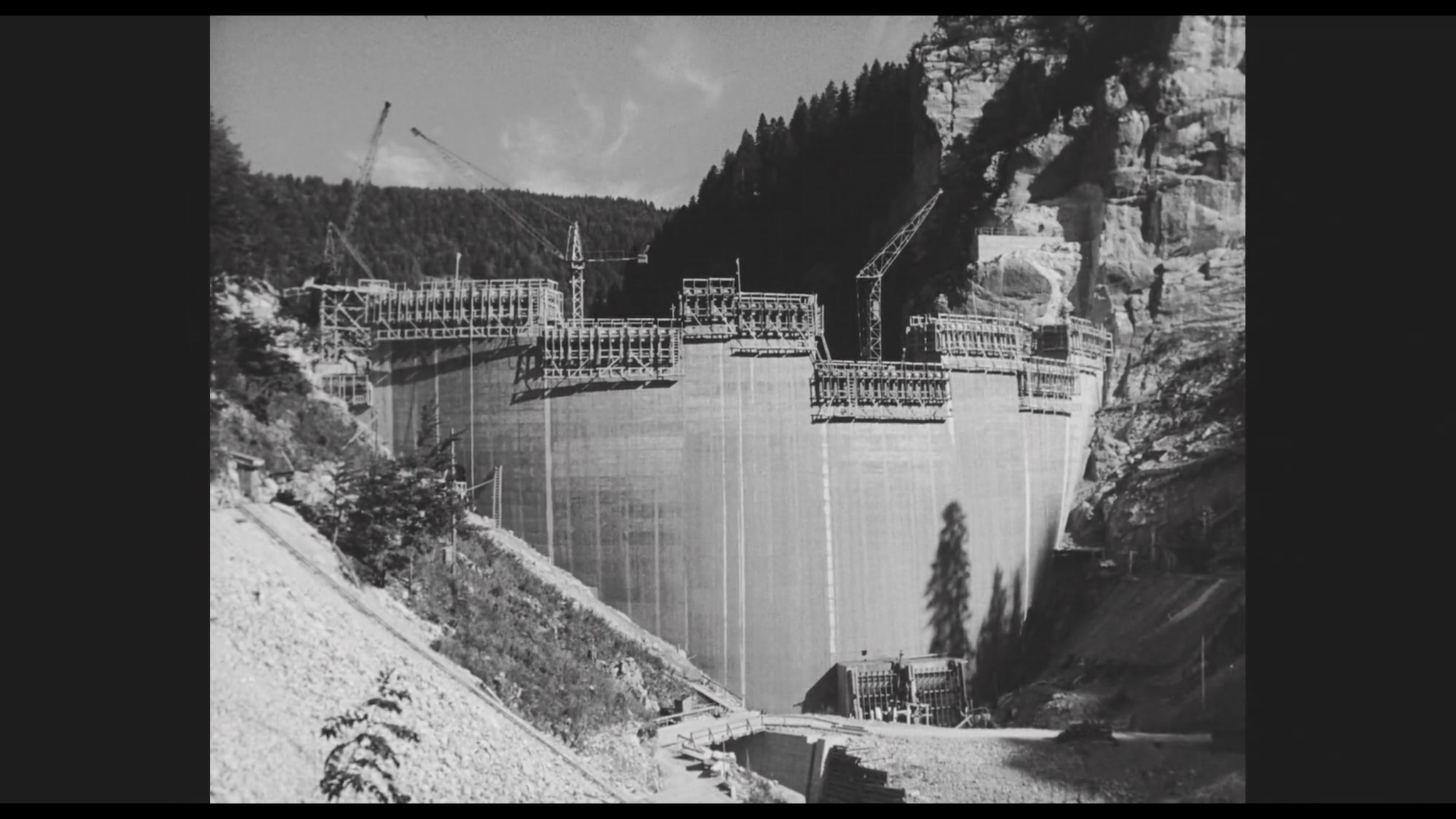Black and white scene of the construction of a large dam in a mountainous area, with cranes and scaffolding, surrounded by trees and rocky terrain.