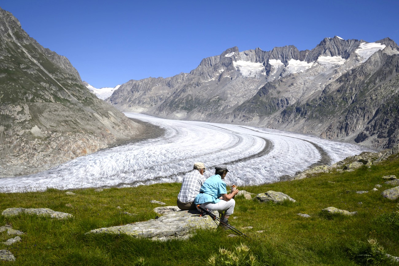 Zwei Menschen sitzen auf einem grossen Stein, im Hintergrund ein grosser Gletscher und Berge