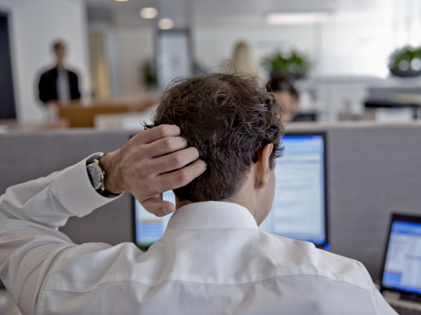 uomo di spalle davanti a pc si gratta la testa con la mano sinistra
