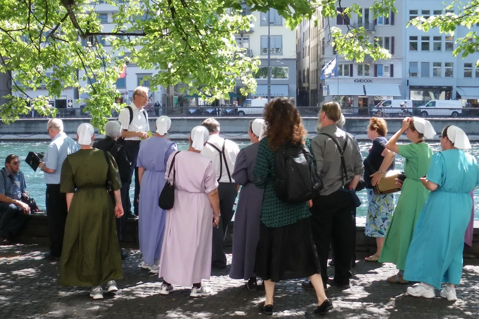 Frauen mit weissen Hauben und Männer mit Hosenträgern stehen an einem Fluss