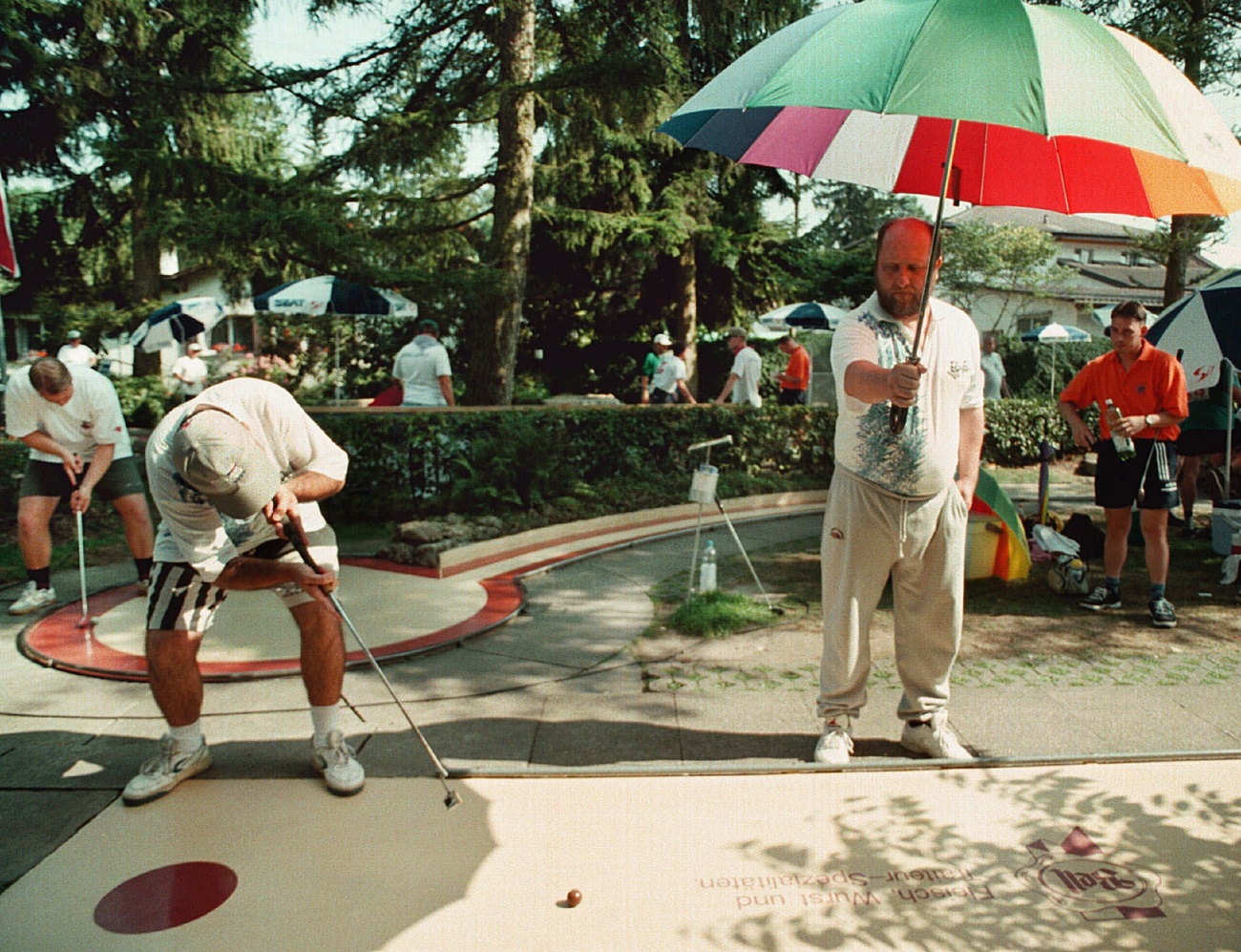 Mehrere Männer auf einer Minigolf-Anlage, ein Mann macht dem Spieler mit einem Schirm Schatten