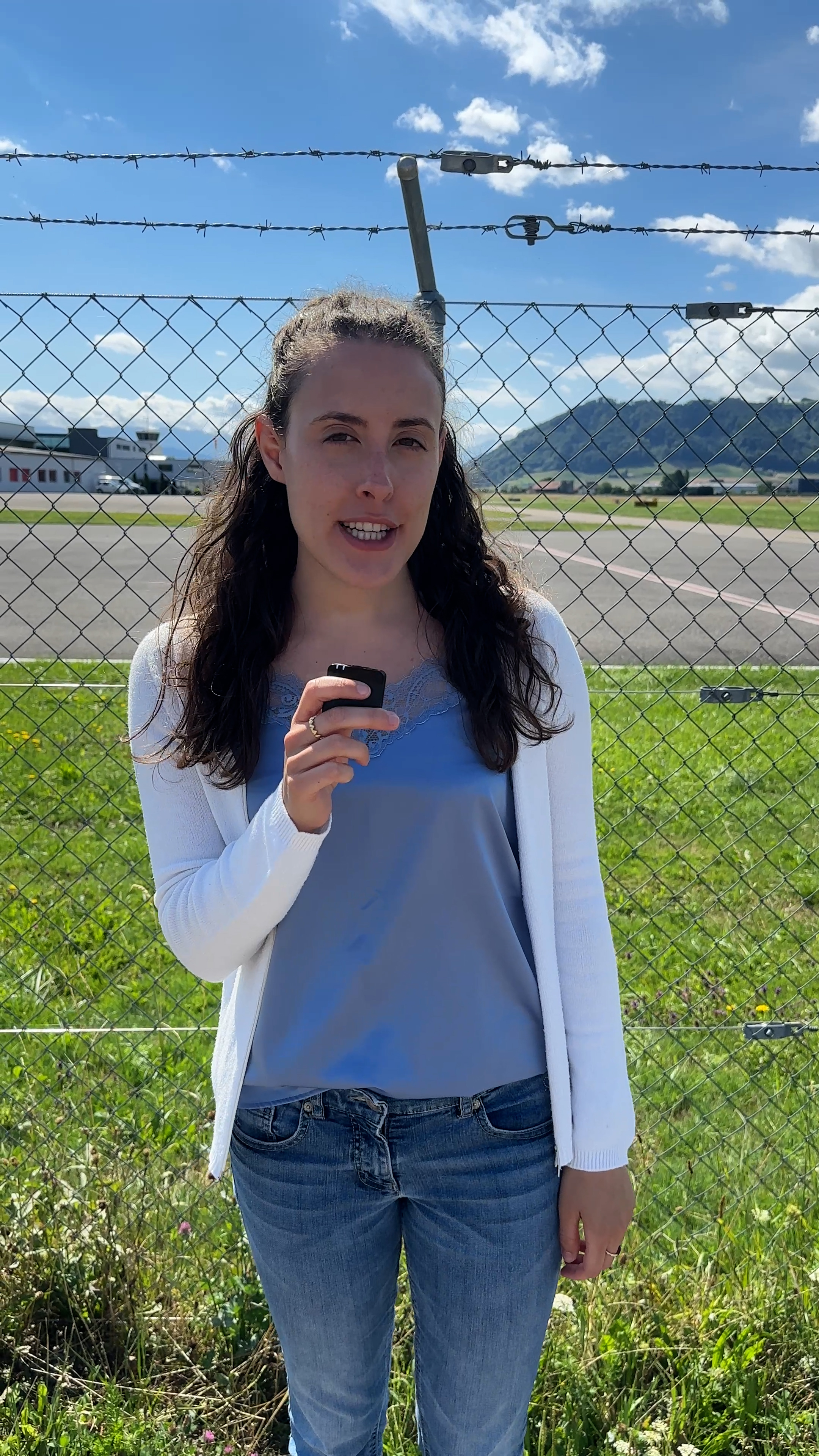 A woman speaking into a microphone with an airport in the background.