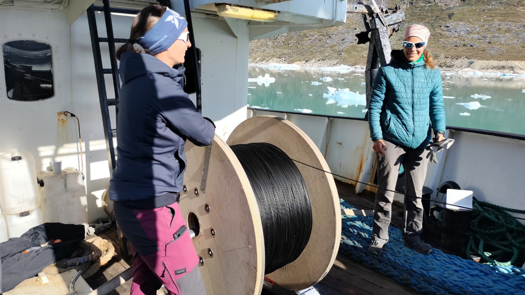 Zwei Frauen auf einem Schiff lassen ein Glasfaserkabel von einer Spule bis zum Meeresboden hinab