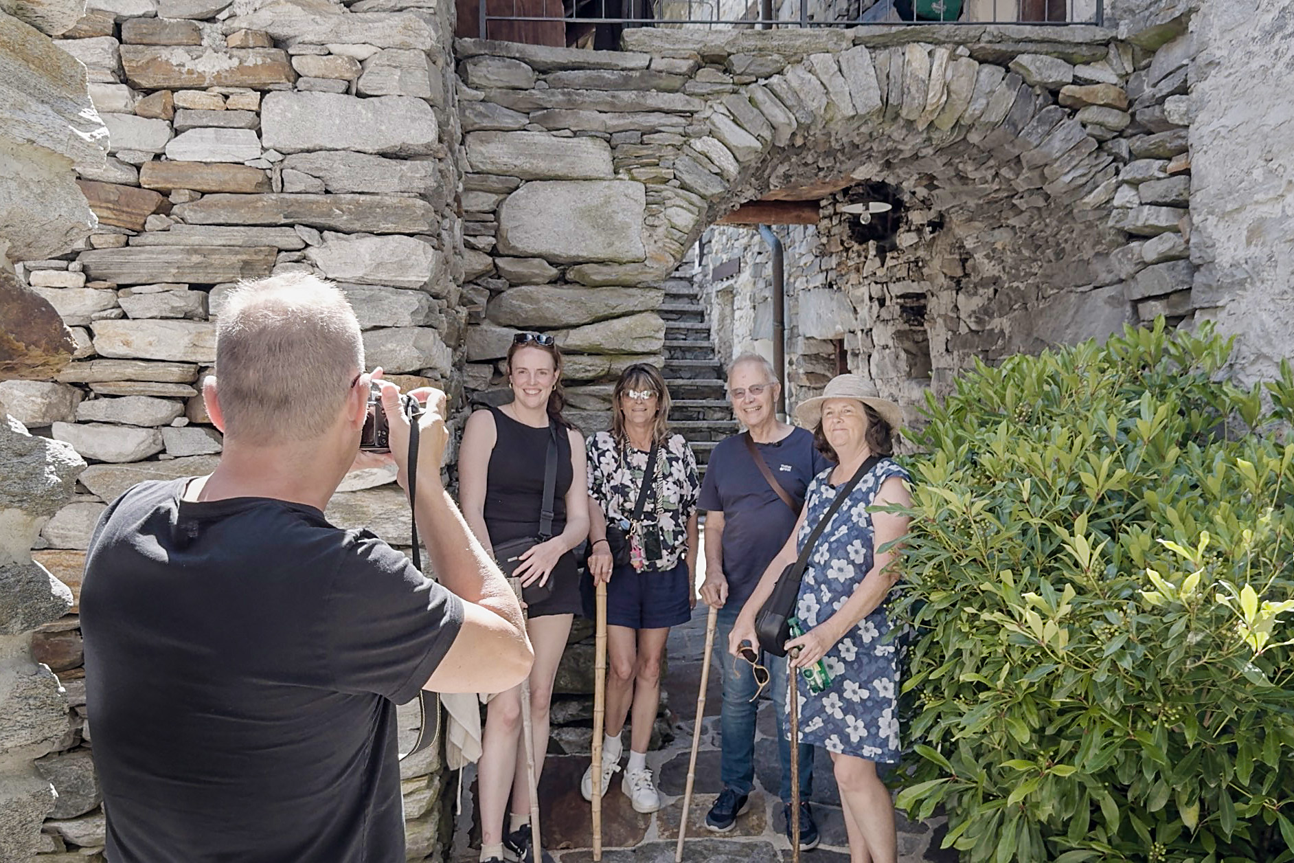 A man photographing a group of 3 women and 1 man standing in front of stone houses