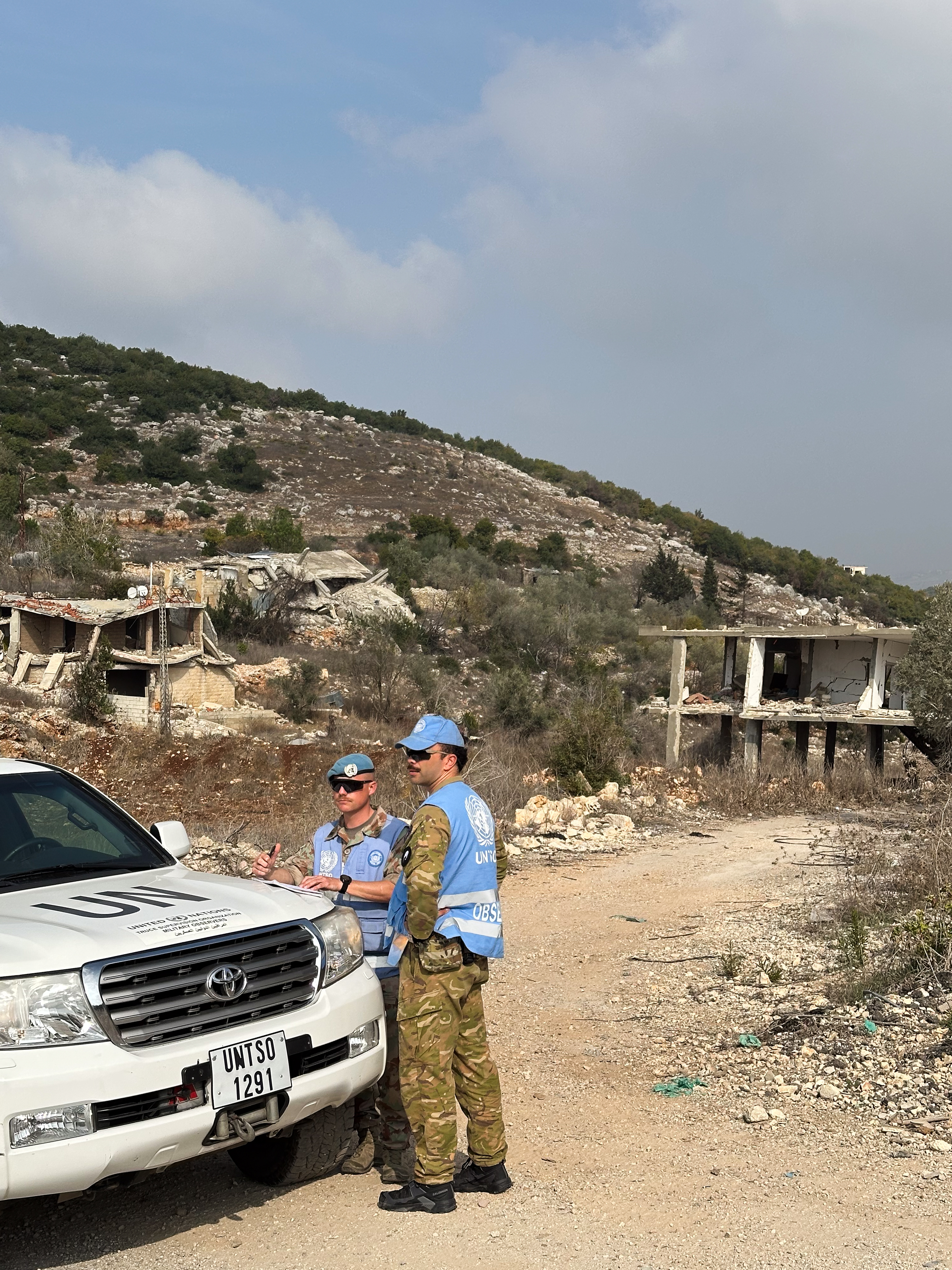 Zwei Personen stehen neben einem Fahrzeug, im Hintergrund zerstörte Gebäude