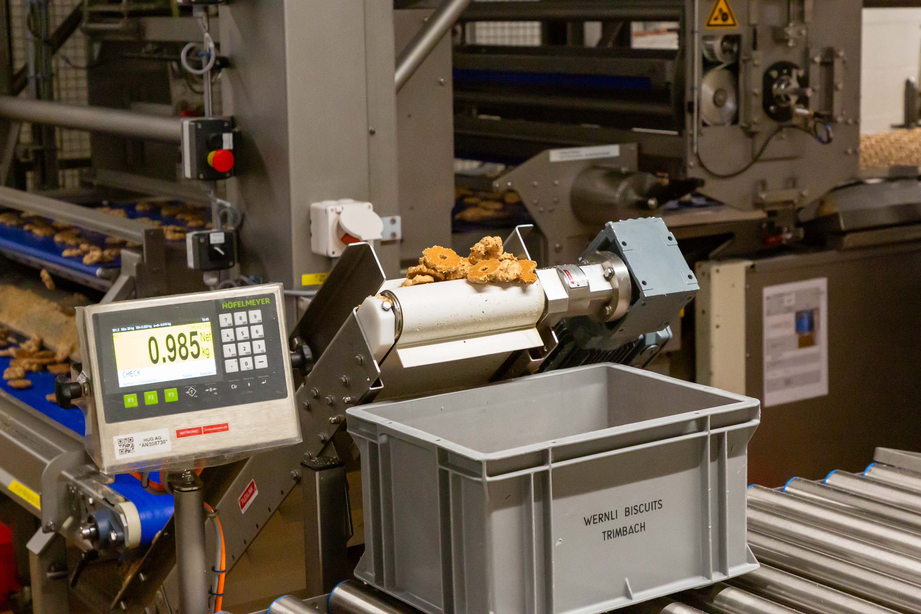 A conveyor system shows a scale measuring the weight of cookies that fall into a gray container labeled "Wernli Biscuits Trimbach"