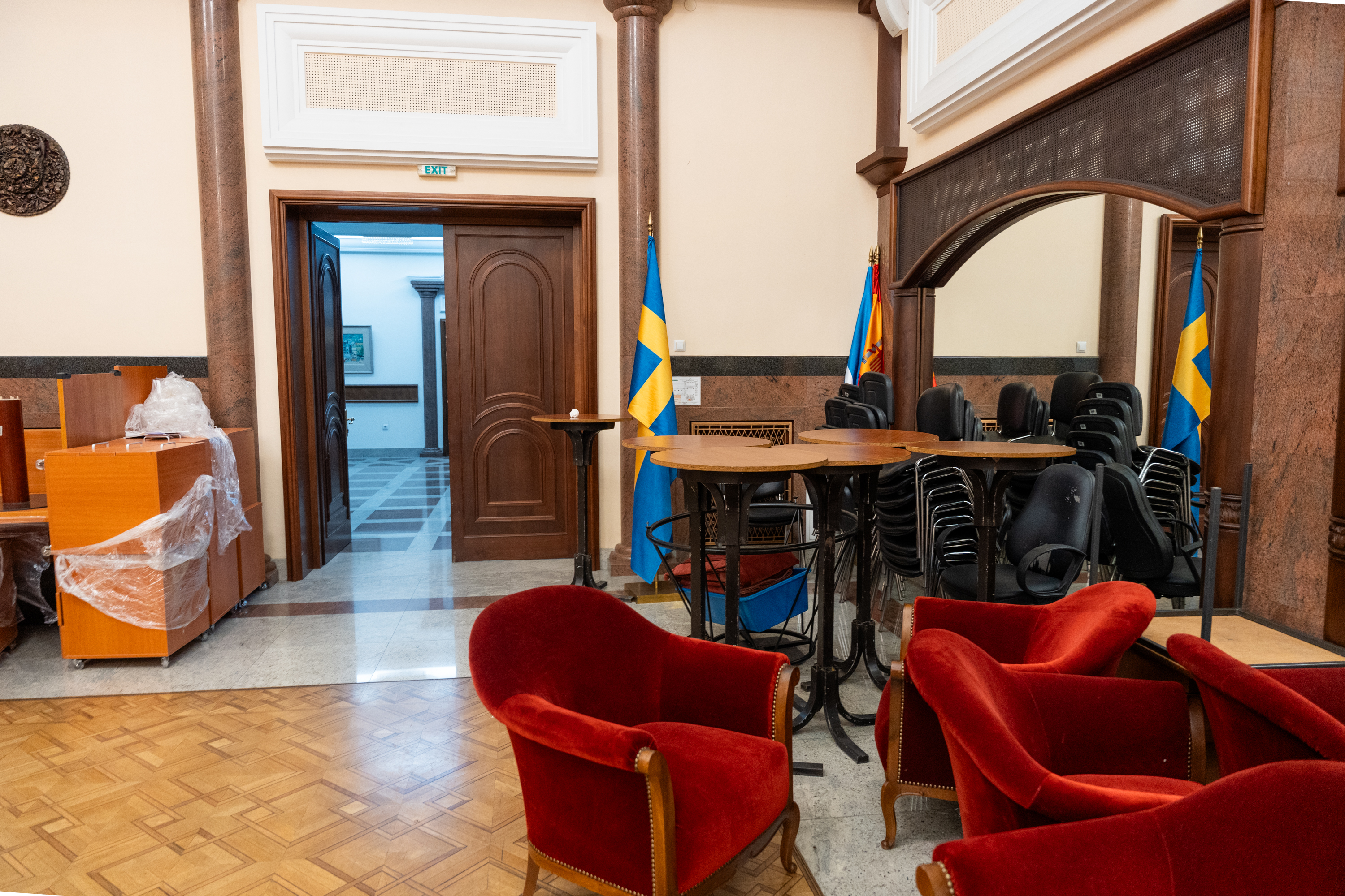 Une pièce avec des chaises en velours rouge, des chaises empilables noires, des tables et des drapeaux, avec une porte en bois menant à une autre zone.