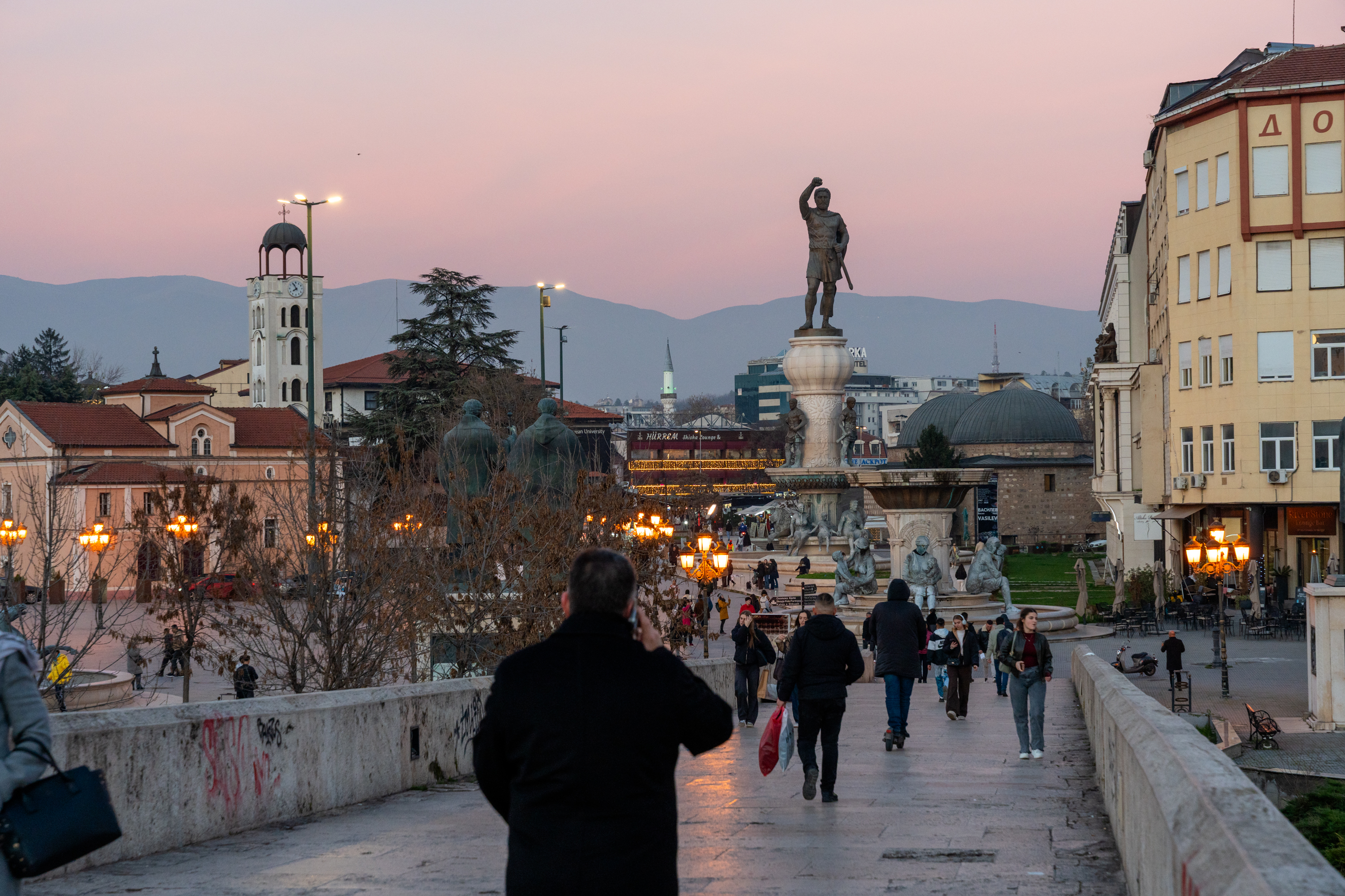 A busy square at dusk with a large statue and people on the pavement