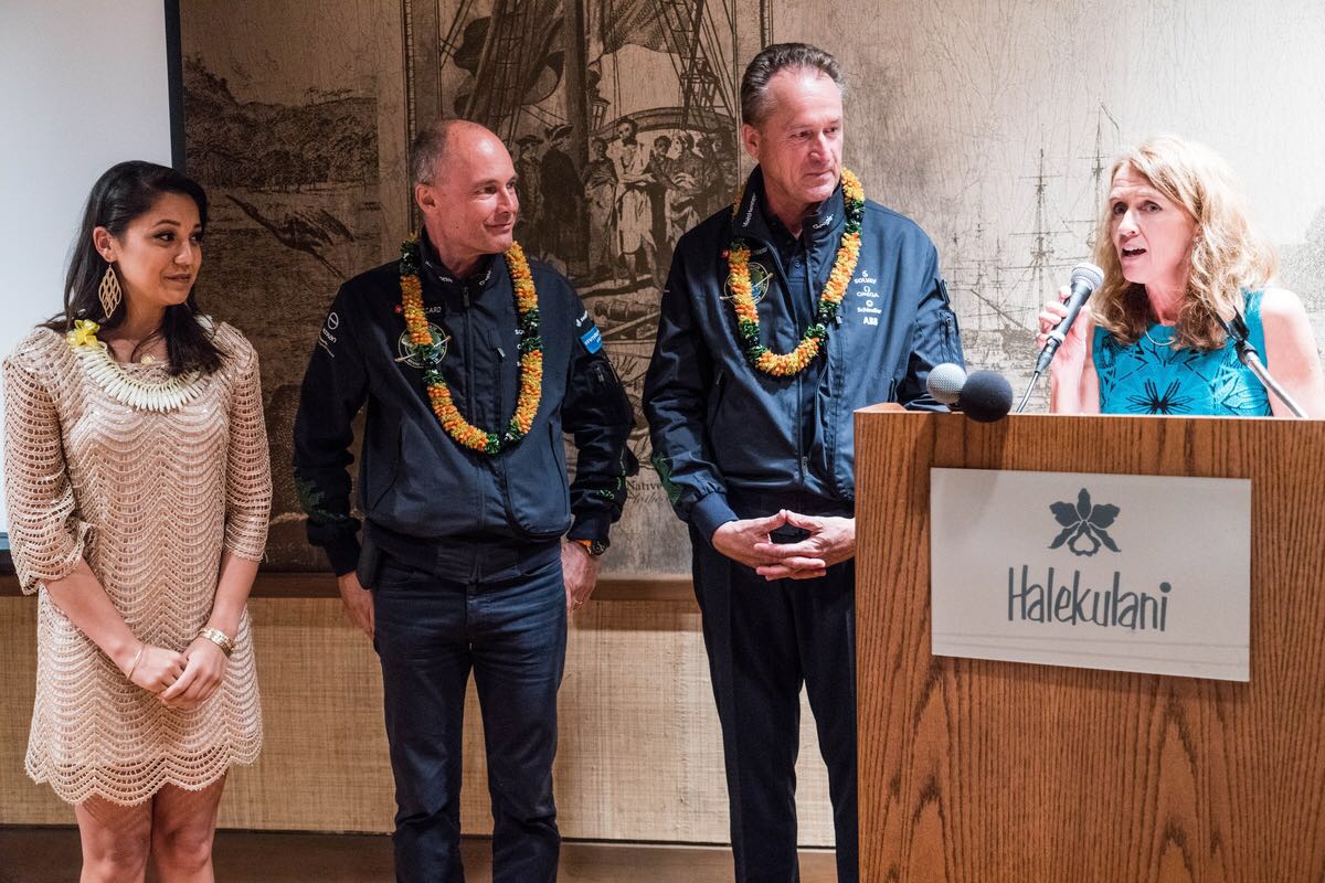 Deux femmes et deux hommes, tous portant des guirlandes de fleurs autour du cou, l'une des femmes prononce un discours.