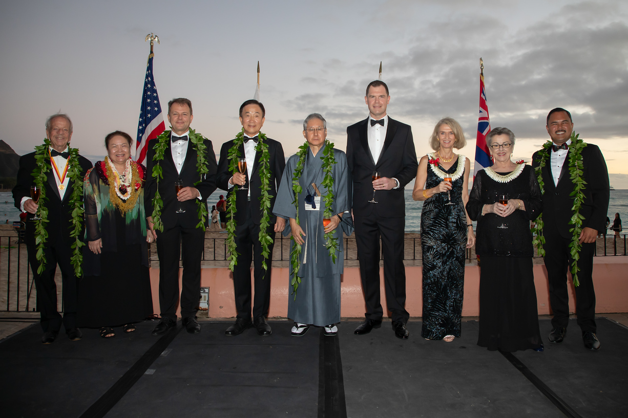 Des dames et messieurs en tenue de cérémonie sont alignés, derrière eux des drapeaux de différents pays.