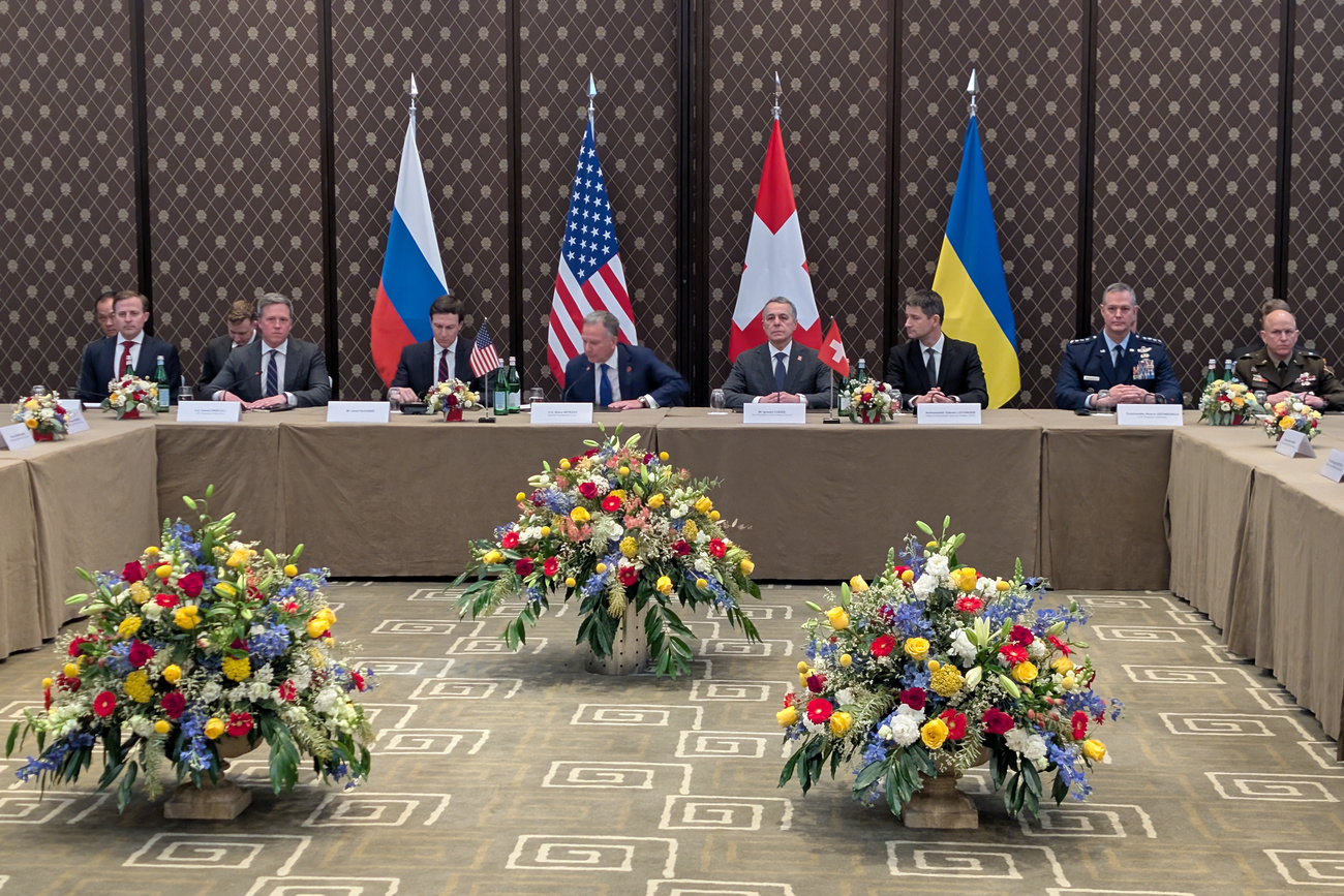 Photo of people at conference table with Russian, US, Swiss and Ukrainian flags in the background