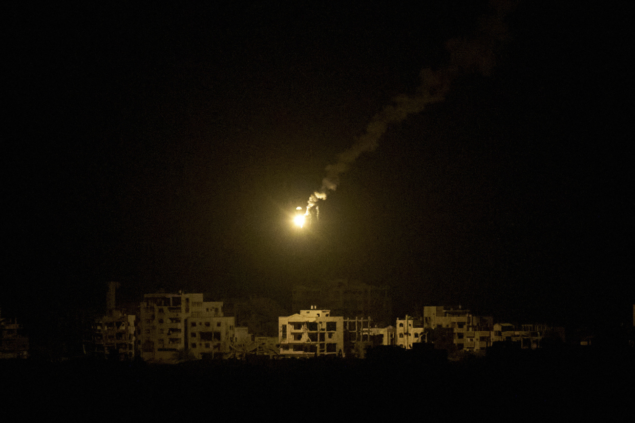 Army flare streaks across dark night sky above silhouetted buildings