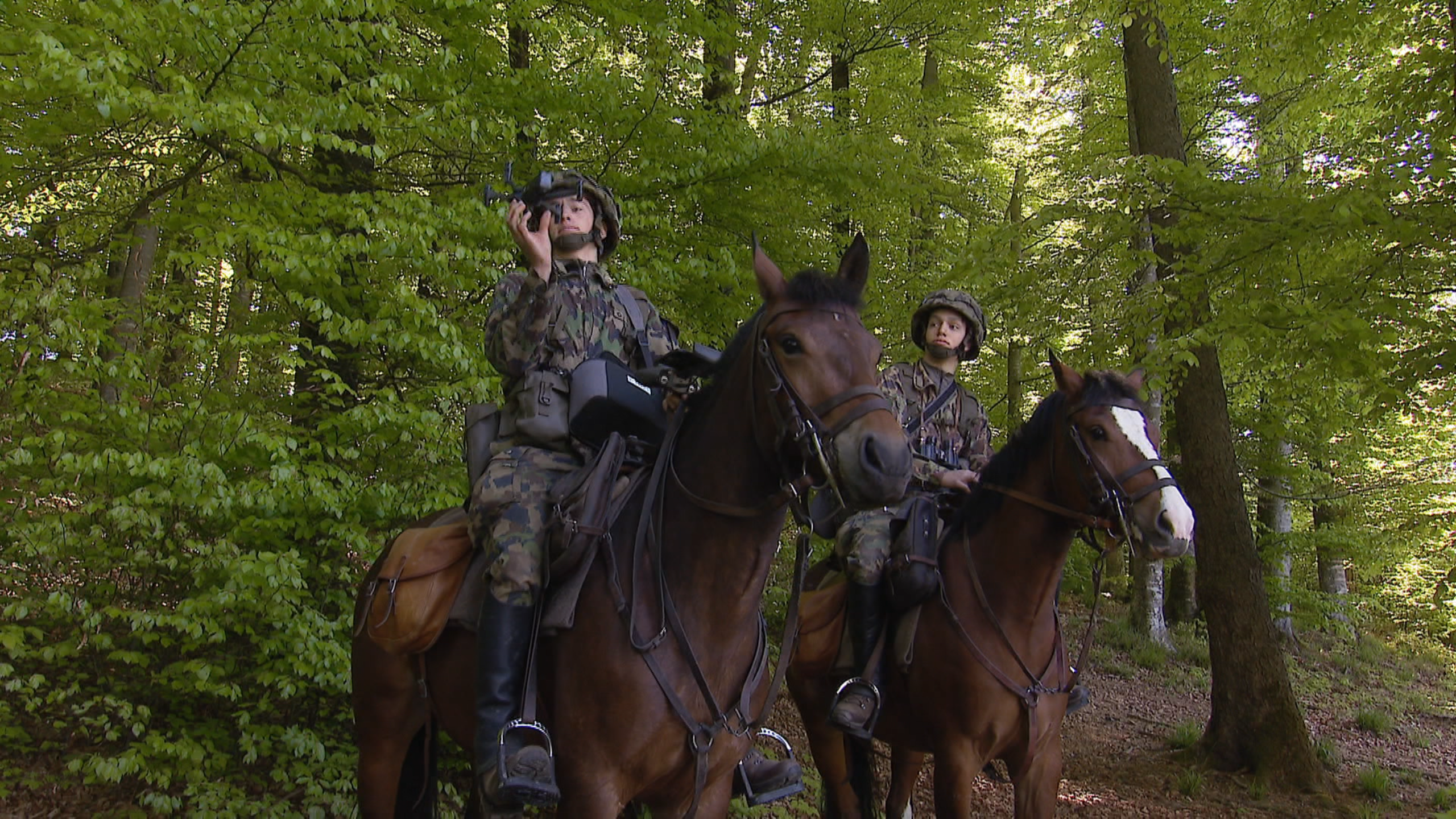 Two people in military uniform on horseback, one of whom is holding a drone aloft