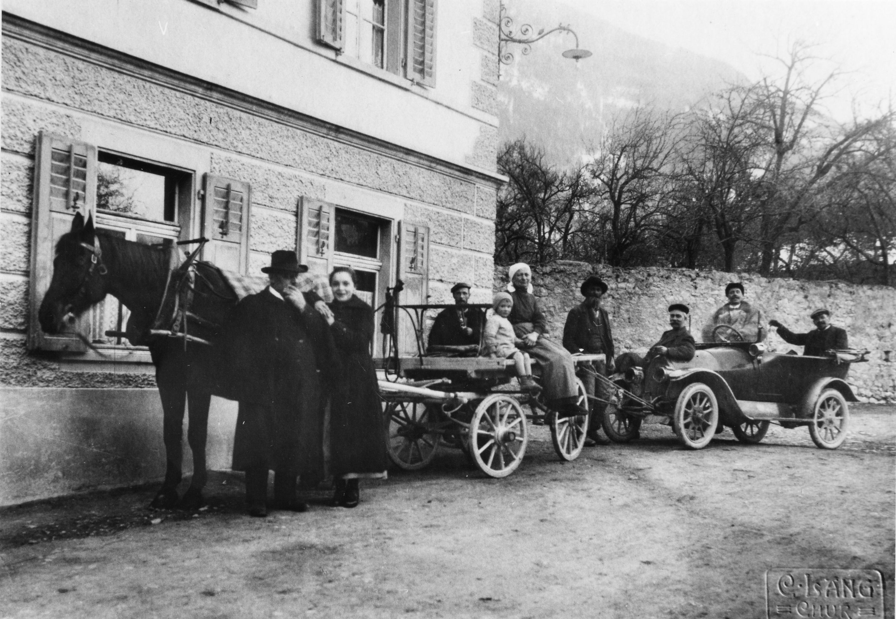 Ein offenes Automobil wird von einem Pferd und einem Pferdewagen dazwischen abgeschleppt. Mehrere Menschein sitzen auf den Gefährten, zwei stehen beim Pferd. Das Bild entstand zwischen 1900 und 1911