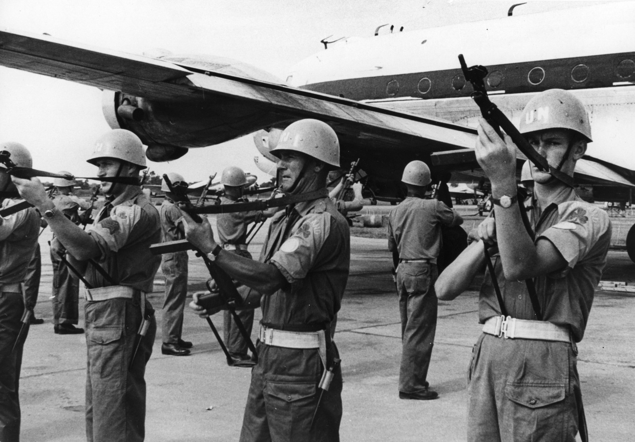 UN soldiers in uniform, holding rifles, stand in formation on an airfield, with a aircraft in the background