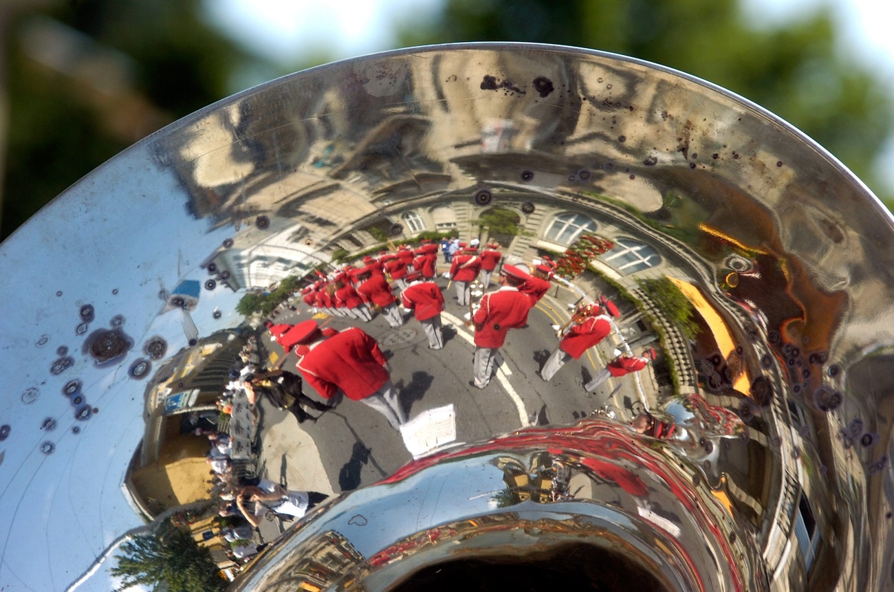 Reflexion von Musikanten in einem glänzenden, zerkratzten Tuba-Schalltrichter, deren Mitglieder rote Jacken und weiße Hosen tragen und sich im Freien befinden.