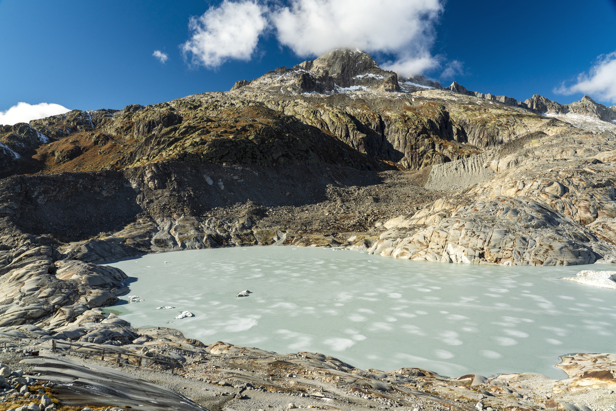A mountain landscape with a glacial lake in the foreground. The water is icy and pale, and surrounded by rocky terrain.