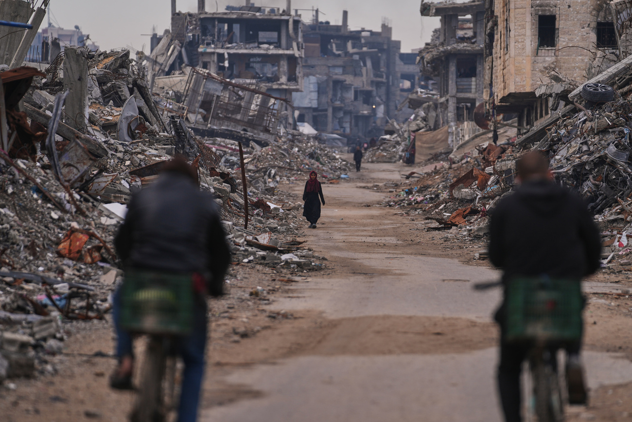 Two people on bicycles ride along a street littered with rubble and lined with damaged buildings, whilst in the distance a person is walking