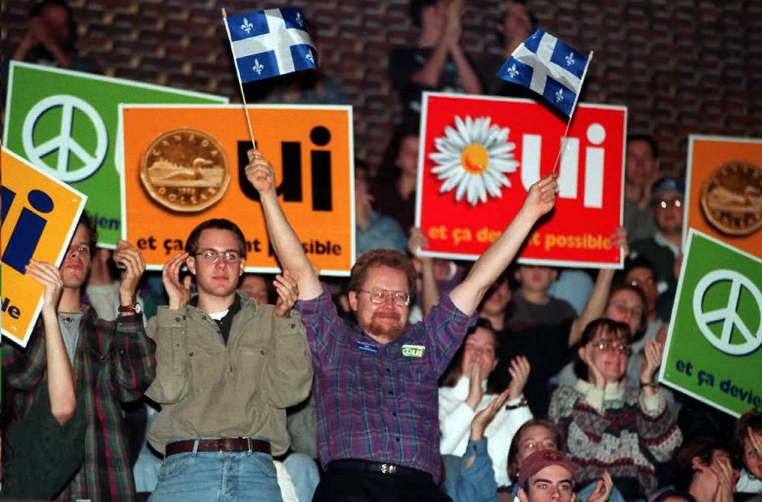 People at a rally; one person is holding up Quebec flags and there are colourful signs bearing the word "oui".
