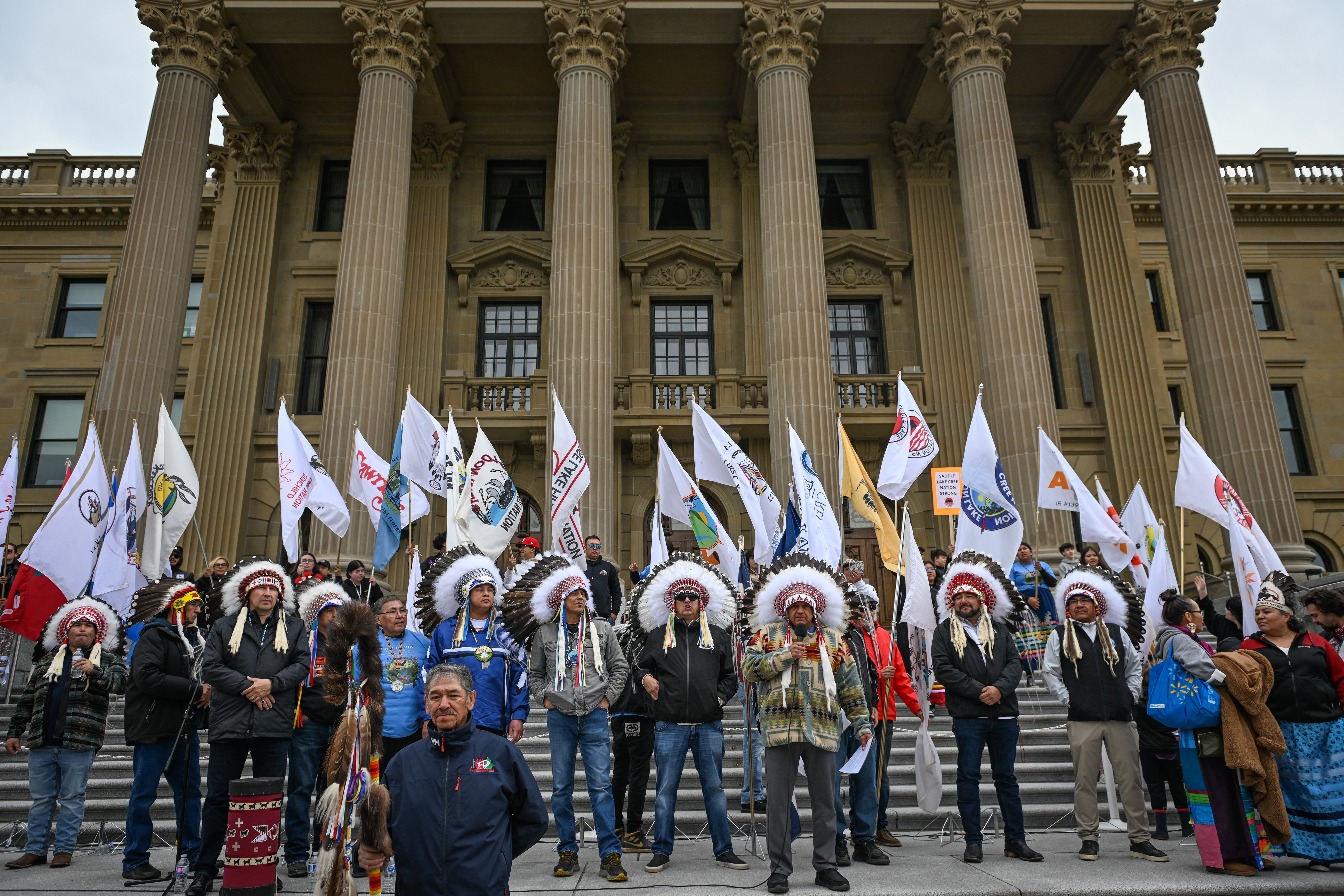 A group of people wearing traditional headgear are standing on the steps of a large, columned building, holding various flags