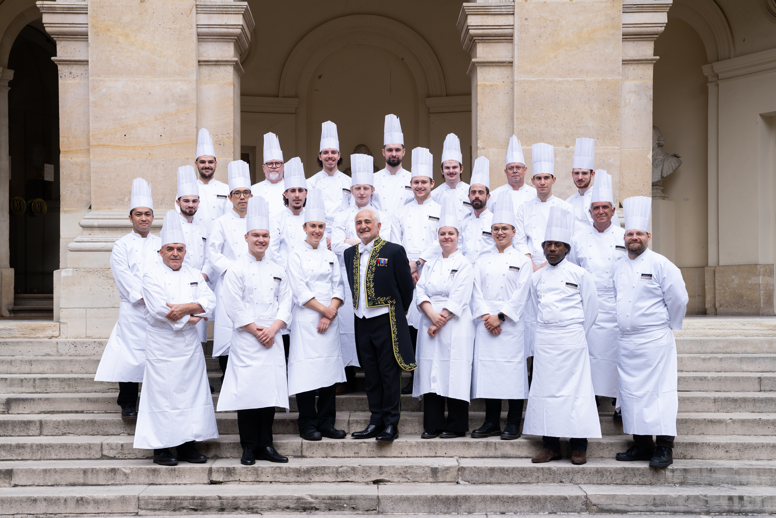 Un groupe de personnes, toutes entièrement vêtues de blanc et coiffées d'un chapeau, au milieu duquel se tient une personne vêtue différemment