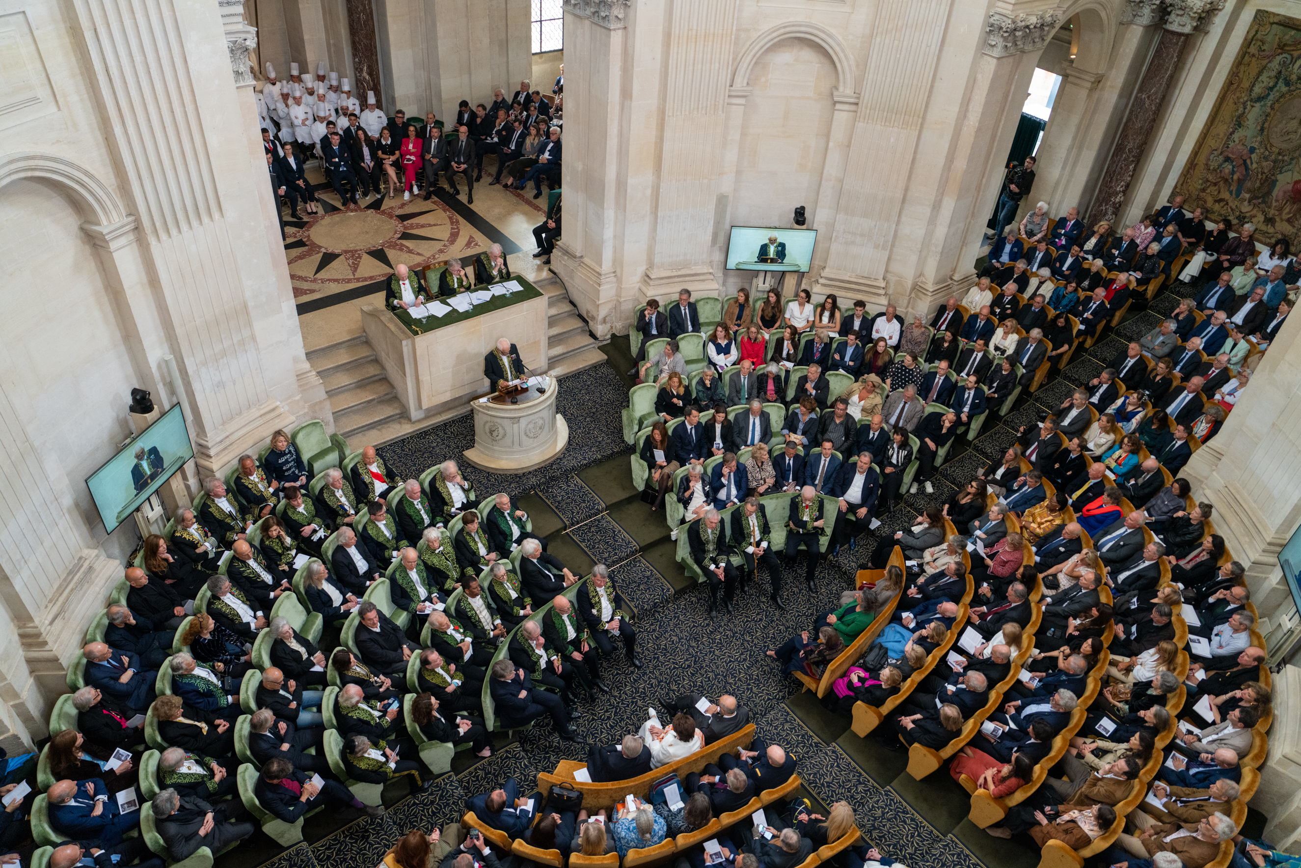 Vue aérienne d'une cérémonie officielle dans une grande salle. Les participants, en costume et en tenue de soirée, sont assis en rangées incurvées et écoutent un orateur depuis un podium.