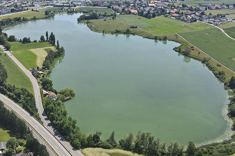 Vue aérienne d'un lac entouré de champs verdoyants, de routes, d'arbres et d'une zone périphérique avec des maisons