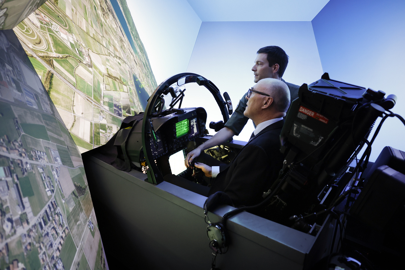 Ein älterer Mann im Anzug sitzt im Cockpit eines Kampfjet-Simulators, er befindet sich im Sturzflug und wird von einem jüngeren Mann assistiert
