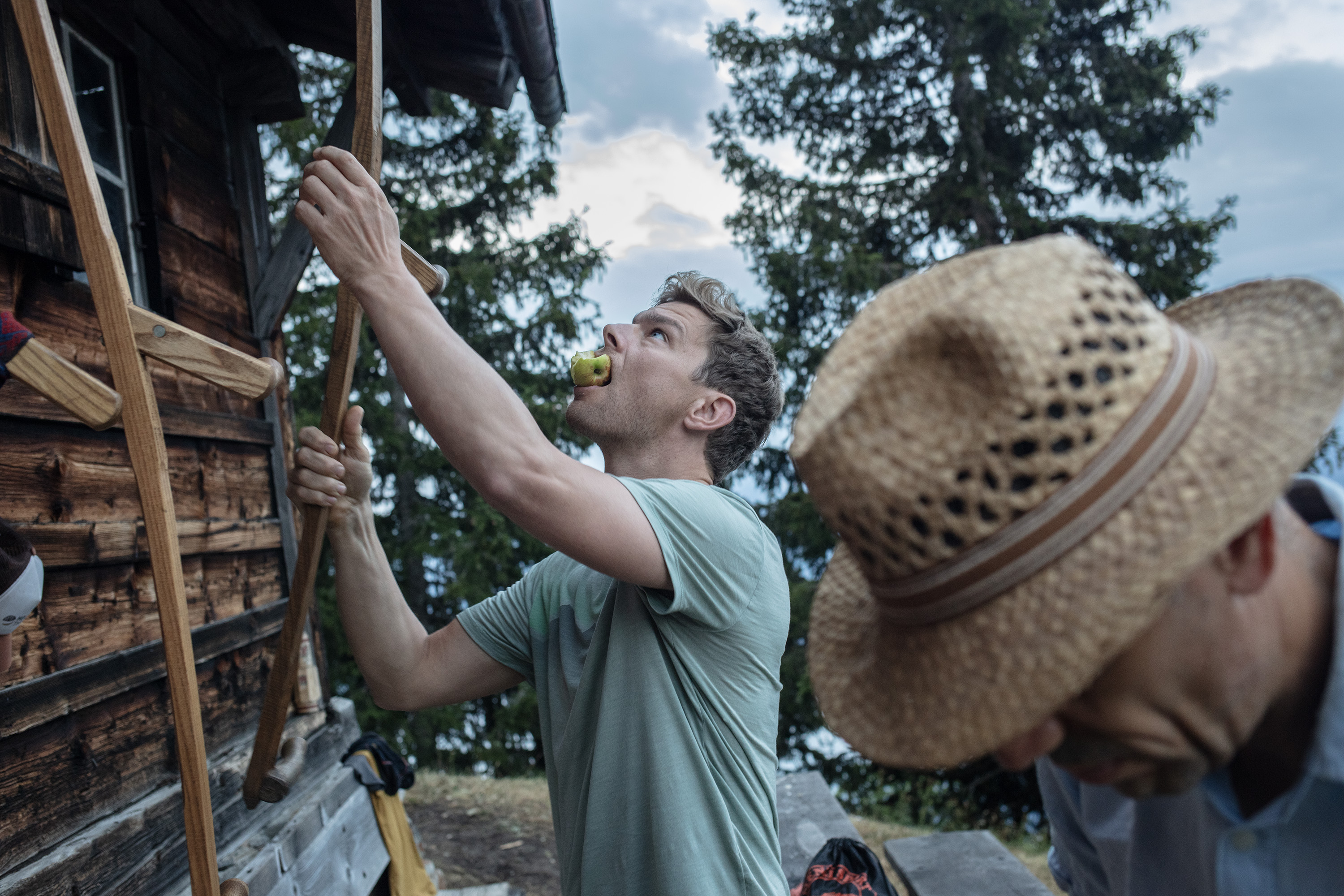 Zwei Männer vor einer Berghütte aus Holz. Einer hängt eine Sense auf und hat einen Apfel im Mund, der andere trägt einen Strohhut