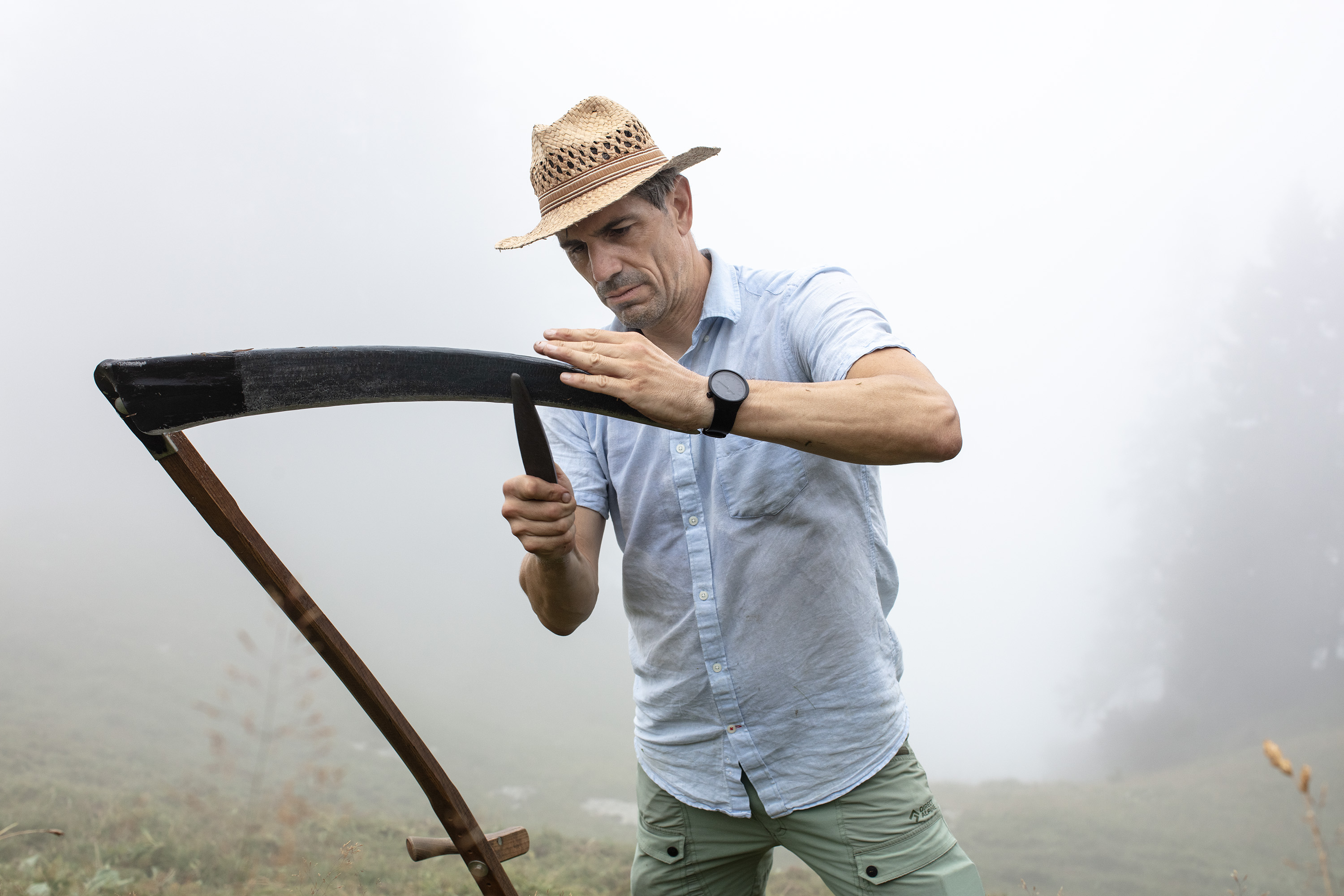 Ein Mann mit Strohhut schärft im Nebel eine Sense