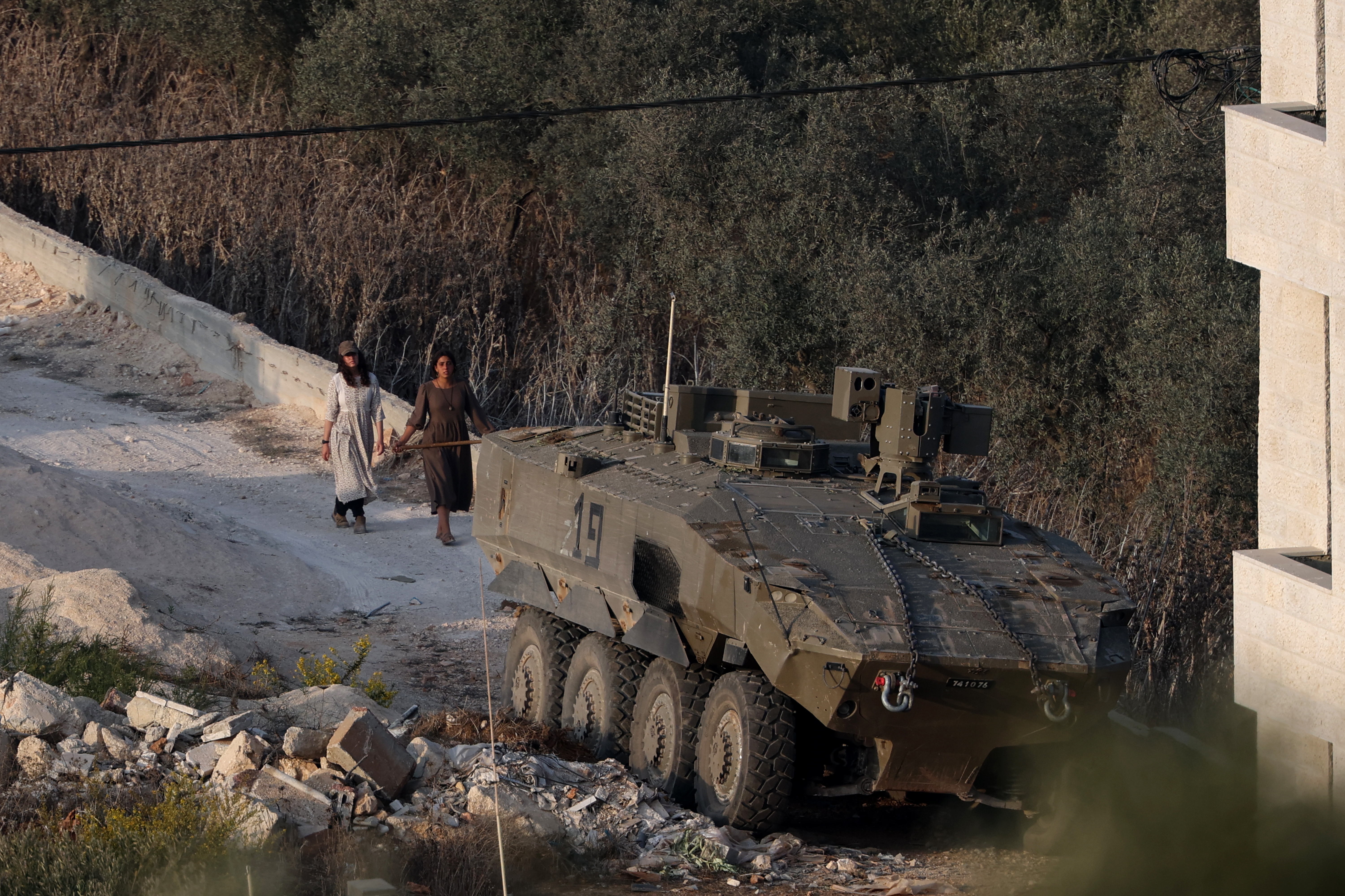 Israeli settlers from a nearby outpost walk past an Israeli military Armored Personnel Carrier (APC) stationed near an occupied building during the annual olive harvest in the Palestinian town of Turmus Ayya, in the West Bank, 24 October 2026.