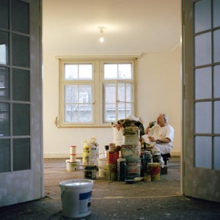 Maler trinkt sitzend neben gestapelten Farbeimern.