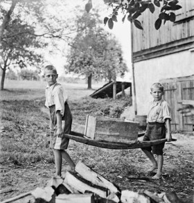 Two boys at a farm carrying a wooden box with a barrow