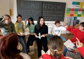 Kinder und eine Lehrerin in einem Klassenraum. An der Wandtafel stehen Sätze in Rätoromanisch.